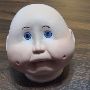 May include: A vintage doll head with a pale complexion, blue eyes, and a slightly open mouth. The doll head has painted eyebrows and rosy cheeks, with a small nose and a rounded chin. The head is made of a smooth material and is likely porcelain or ceramic.