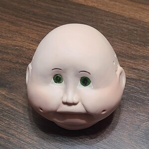 May include: A doll head with a pale complexion, green eyes, and rosy cheeks. The doll head has a rounded shape with small ears and a slightly pursed mouth. The eyebrows are thin and dark, and the head appears to be made of a smooth material.