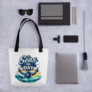 May include: White tote bag with black handles. It features a nautical design with an anchor, waves, and the text "Seas the Day." The design includes blue, orange, and teal colours. Other items in the image include sunglasses, a notebook, and a laptop.