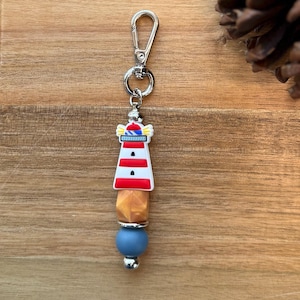May include: A lighthouse-themed keyring with a silver clasp. The lighthouse charm is white with red stripes and a blue and yellow top. Below the lighthouse are a gold-coloured bead and a blue bead.