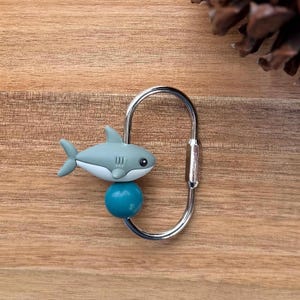 May include: A shark-themed keychain featuring a gray and white shark charm atop a teal blue sphere. The shark is attached to a silver-colored carabiner clip, perfect for keys or bags. The background is a wooden surface.