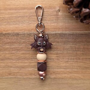 May include: A brown bat-shaped keychain charm with a gold-colored clasp. The charm features a cute bat figure with large eyes, wings, and a small belly. The charm is attached to a beaded design with brown, beige, and rose gold beads.