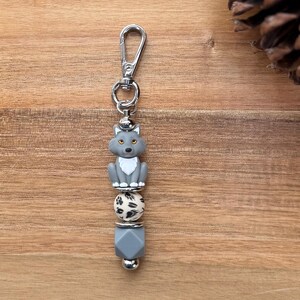 May include: A silver keychain with a gray wolf charm, a black and white patterned bead, and a gray geometric bead. The wolf has yellow eyes and a white chest. The keychain has a silver clasp.