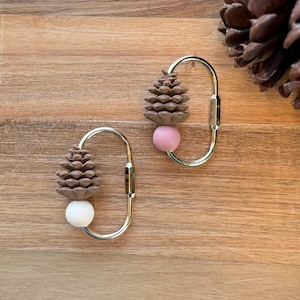 May include: Two gold-colored carabiner clips, each adorned with a brown pinecone, a pink or white sphere, and a silver clasp. The clips are set against a wooden background, with a pinecone in the upper right corner.