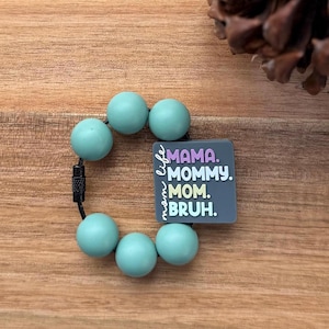 May include: A teal teething ring with six round beads and a black cord. A gray square charm features the words "MAMA. MOMMY. MOM. BRUH." in white text. The charm also has the words "mom life" in a script font.