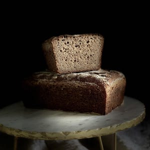 Könnte beinhalten: Zwei frisch gebackene Brote, eines ganz und eines geschnitten, sind auf einem Tisch mit Marmorplatte und goldenen Beinen gestapelt. Das Brot hat eine dunkelbraune Kruste und ein hellbraunes Inneres, vor schwarzem Hintergrund.