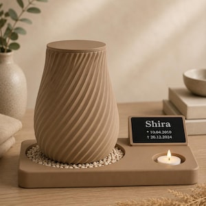Könnte beinhalten: Eine beige Urne mit spiralförmig texturierter Oberfläche steht auf einem rechteckigen Sockel, der mit kleinen Steinen gefüllt ist. Neben der Urne befindet sich eine schwarze Plakette mit dem Text Shira, 10.04.2010, 20.12.2024 und ein brennendes Teelicht.