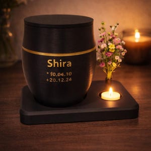 Könnte beinhalten: Schwarze Urne mit goldenem Band und dem Namen "Shira" in goldener Schrift. Die Daten 10.04.10 und 20.12.24 stehen darunter. Die Urne steht auf einem schwarzen quadratischen Sockel mit einem kleinen Kerzenhalter und einer brennenden Kerze.