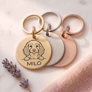 May include: Three circular metal pet tags with split rings, featuring a gold, silver, and rose gold finish. The gold tag is engraved with a line art illustration of a curly-haired dog and the name MILO printed in black capital letters.
