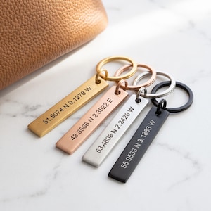 May include: Four rectangular metal keychains in gold, rose gold, silver, and black finishes. Each keychain features a circular ring and is engraved with specific geographic coordinates: 51.5074 N 0.1278 W, 48.8566 N 2.3522 E, 53.4808 N 2.2426 W, and 55.9533 N 3.1883 W.