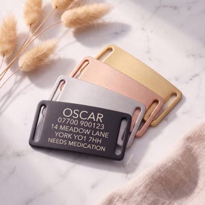 May include: A set of four rectangular metal pet ID tags with rounded corners and side slots for collar attachment. The tags are finished in black, silver, rose gold, and gold. The black tag is engraved with the text OSCAR, 07700 900123, 14 MEADOW LANE, YORK YO1 7HH, NEEDS MEDICATION.