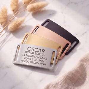 May include: Four rectangular metal pet ID tags with rounded corners and side slots for collar attachment. The tags are finished in silver, gold, rose gold, and black. The silver tag is engraved with the text OSCAR, 07700 900123, 14 MEADOW LANE, YORK YO1 7HH, NEEDS MEDICATION.