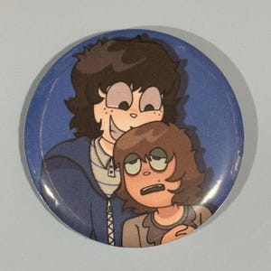 May include: A round button with a blue background featuring an illustration of two cartoon characters. One character has dark hair and a mischievous grin, while the other has brown hair and a concerned expression. The button is a collectible item.