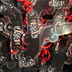 Op de afbeelding: Transparante acryl sleutelhanger met een stripfiguur met kattenoren, gekleed in een camouflage shirt en een zwarte broek. De sleutelhanger heeft een rode sluiting. Het ontwerp wordt meerdere keren herhaald.