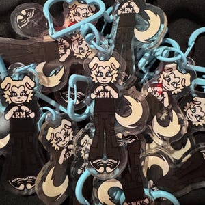 May include: A collection of clear acrylic keychains featuring a cartoon character with a white face, black outfit, and the letters "ARM." The keychains have light blue clasps and a crescent moon design. The background is dark.