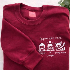 May include: A burgundy sweatshirt featuring the French phrase "Apprendre c'est essayer, se tromper, progresser" in white. The design includes three cartoon figures and a rising graph, promoting learning and growth.