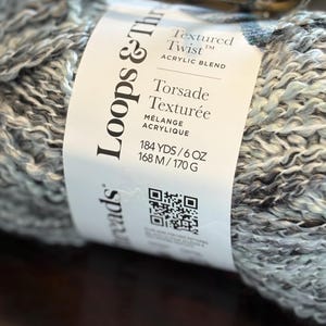 Puede incluir: Un ovillo de hilo acrílico mezclado Loops & Threads Textured Twist. La etiqueta dice "Torsade Texturée" e incluye medidas de 168 m/170 g. El hilo es una mezcla de gris, blanco y azul claro.