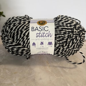 May include: A skein of Basic Stitch yarn in black and white. The yarn is tightly wound and has a striped appearance. The label reads "Basic Stitch" and "anti-pilling". The yarn is suitable for knitting projects.
