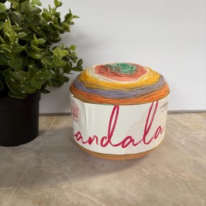 May include: A vibrant ball of yarn in shades of yellow, orange, green, and purple. The yarn is wrapped around a label with the word "mandala" in pink script. A small green plant is visible in the background. Ideal for knitting and crochet projects.