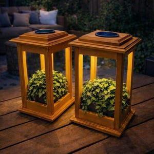 May include: Two wooden lanterns with a warm, yellow glow. Each lantern has a square top with a dark blue circle and a base. Inside each lantern is a green plant. The lanterns are on a wooden surface.