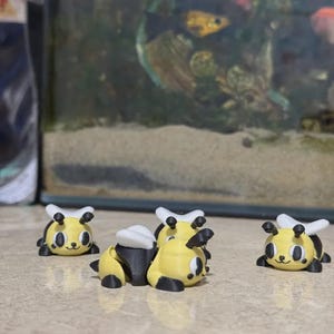Könnte beinhalten: Eine Sammlung gelb-schwarzer Bienenfiguren mit weißen Flügeln und schwarzen Antennen. Die Figuren haben lächelnde Gesichter und sind auf einer hellen Oberfläche angeordnet. Im Hintergrund ist ein kleines Aquarium zu sehen.