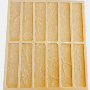 Puede incluir: Un molde rectangular de silicona con doce cavidades verticales estrechas. Cada cavidad tiene una superficie texturizada e irregular que recuerda a la piedra natural o a la pizarra. El molde es de color beige claro y tiene una estructura de rejilla.