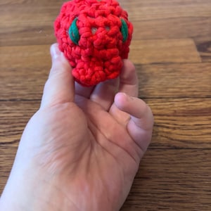 May include: A handmade, red crocheted ball with two green leaf-shaped accents. The ball is held in a hand against a wooden background. The ball's texture is visible, showcasing the tight crochet stitches.