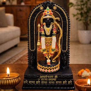 May include: A black statue of a deity holding a golden bow and arrow, adorned with colorful jewels and white beads. The base features gold Hindi text that reads Ayodhya ke Ram Lalla and Hare Rama Hare Rama Rama Rama Hare Hare.
