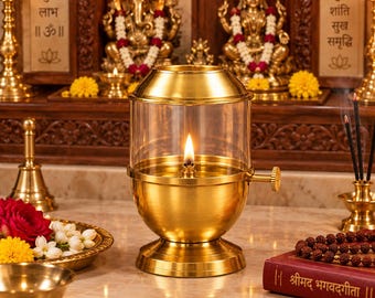 May include: A brass oil lamp with a clear glass chimney and a side adjustment knob. The lamp features a polished gold-tone finish, a circular base, and a lit wick centered inside the glass enclosure, providing a steady flame for illumination.