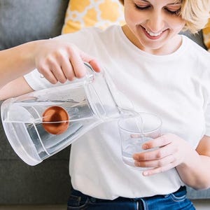 May include: A clear glass pitcher with a round terracotta-colored insert inside, being used to pour water into a clear glass tumbler. The pitcher features a sturdy handle and a spout, while the terracotta insert sits submerged within the vessel.