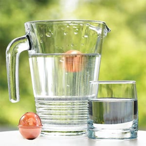 May include: A clear glass pitcher filled with water and a single copper-colored spherical object submerged inside. A matching copper-colored sphere sits on the surface next to a clear glass tumbler also filled with water, reflecting light.