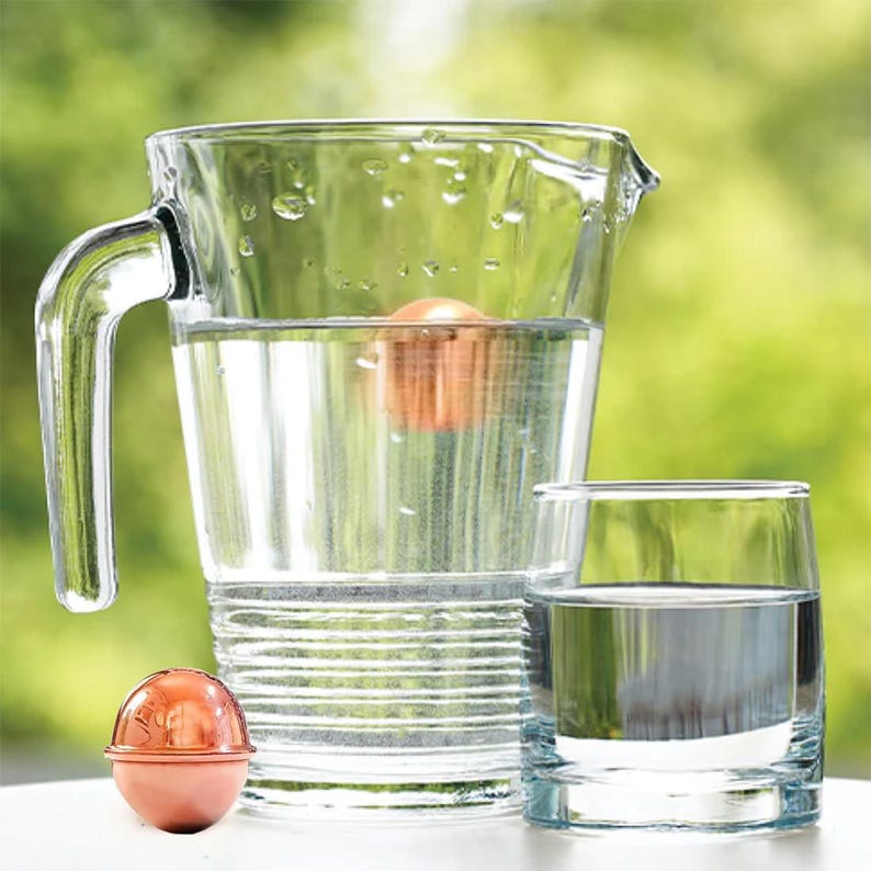 May include: A clear glass pitcher filled with water and a single copper-colored spherical object submerged inside. A matching copper-colored sphere sits on the surface next to a clear glass tumbler also filled with water, reflecting light.