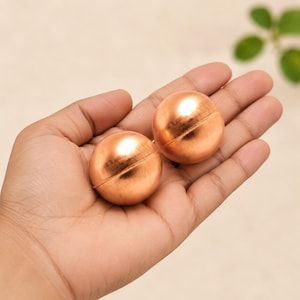 Op de afbeelding: Twee gepolijste koperen bollen met een glad, reflecterend oppervlak en een zichtbare horizontale naad die rond het midden van elke bal loopt. Deze metalen bolvormige objecten hebben een warme, roodachtig oranje kleur en een ronde vorm.