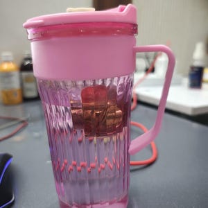 May include: A transparent pink plastic tumbler with vertical ribbed detailing, featuring a matching pink lid with a small white tab and a pink side handle. The cup contains a submerged, brownish-red object visible through the clear walls.