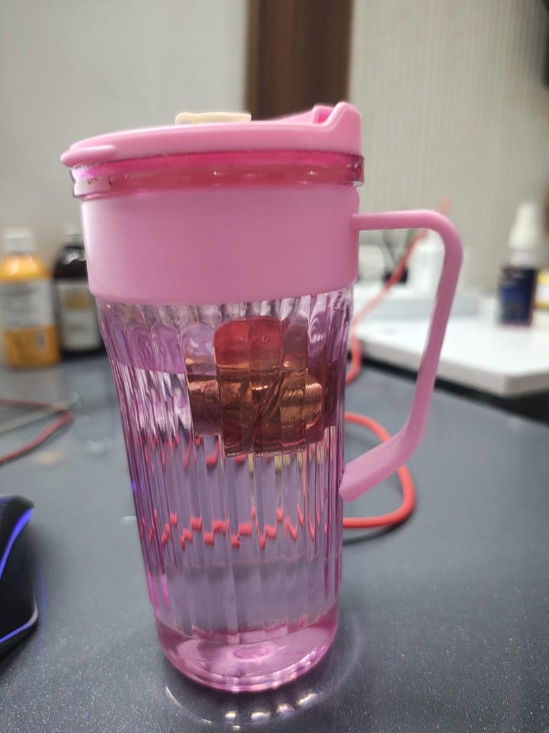 May include: A transparent pink plastic tumbler with vertical ribbed detailing, featuring a matching pink lid with a small white tab and a pink side handle. The cup contains a submerged, brownish-red object visible through the clear walls.