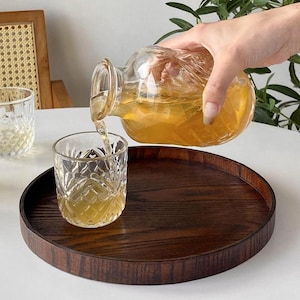 May include: A clear glass carafe with a textured surface pouring amber liquid into a matching patterned glass tumbler. Both items are positioned on a circular dark brown wooden tray with a visible grain, creating a contrast with the clear glassware.
