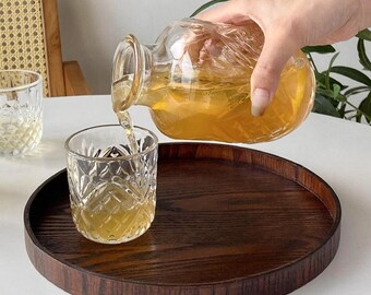 May include: A clear glass carafe with a textured surface pouring amber liquid into a matching patterned glass tumbler. Both items are positioned on a circular dark brown wooden tray with a visible grain, creating a contrast with the clear glassware.
