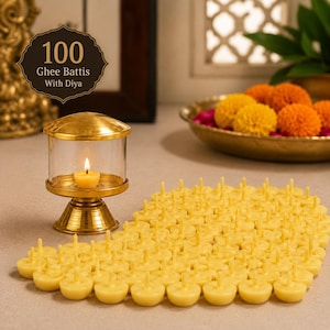 May include: A set of 100 yellow ghee battis arranged on a surface next to a brass diya with a glass cover. The text 100 Ghee Battis With Diya is visible in the upper left corner. The brass diya contains a small lit candle with a yellow flame.