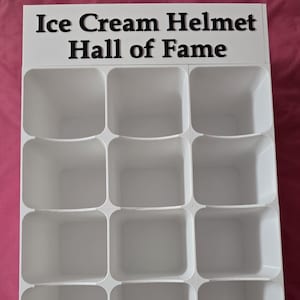 May include: A white display rack featuring a grid of twelve square compartments arranged in four rows and three columns. The top section includes a white header with the black text Ice Cream Helmet Hall of Fame printed in a bold, sans-serif font.