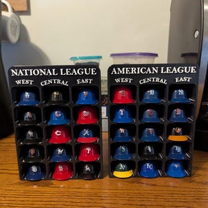 Può includere: Due espositori in plastica nera pieni di cappellini da baseball in miniatura. Uno è etichettato "NATIONAL LEAGUE" e l'altro "AMERICAN LEAGUE". Ogni cappellino rappresenta una squadra, con vari colori e loghi.