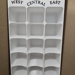 Könnte beinhalten: Ein weißer Kunststoff-Organizer mit fünfzehn quadratischen Fächern, die in drei Spalten angeordnet sind. Die Kopfzeile trägt den schwarzen Schriftzug AMERICAN LEAGUE, mit den Untertiteln WEST, CENTRAL und EAST über jeder Spalte.