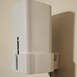 Może przedstawiać: Białe, prostokątne urządzenie zamontowane na ścianie. Urządzenie posiada pionową szczelinę z małymi światełkami i jest połączone z przewodami. Urządzenie to prawdopodobnie modem lub router, przeznaczony do użytku domowego lub biurowego.