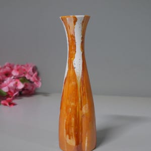Könnte beinhalten: Eine hohe, orangefarbene Keramikvase mit weißem, getropftem Farbmuster. Die Vase hat einen ausgestellten oberen Teil und einen schmalen Boden. Im Hintergrund sind rosa Blumen zu sehen. Die Vase steht auf einer weißen Oberfläche.