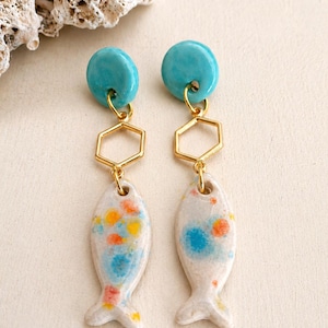 Può includere: Un paio di orecchini a forma di pesce in ceramica. Ogni orecchino presenta un bottone blu turchese, una forma geometrica color oro e un ciondolo a forma di pesce bianco con dettagli blu, gialli e arancioni. Per orecchie forate.