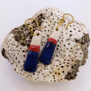 Puede incluir: Un par de pendientes de cerámica hechos a mano con ganchos dorados. Cada pendiente es rectangular, con una parte superior blanca, una sección central roja y una base azul. Se presentan sobre una superficie texturizada de color blanquecino.