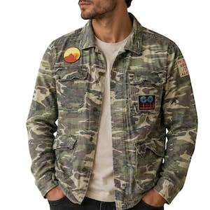 May include: Camouflage print jacket with multiple pockets and patches. The jacket features a desert landscape patch, a patch with two trees, and a patch with text. The jacket is buttoned up and has a collared neckline.