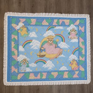 Pode incluir: Um edredom de bebê com fundo azul apresentando um ursinho em uma lua crescente, arco-íris, nuvens e figuras de anjos. O edredom tem uma borda com babados brancos e bordas de patchwork. O texto "MY CHILD'S ANGEL" é impresso na borda.