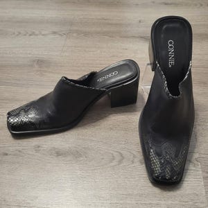 May include: A pair of black leather mules with a square toe and a block heel. The front of each shoe features a textured gray and black snakeskin pattern inlay. The interior footbed is black and displays the brand name CONNIE in white lettering.