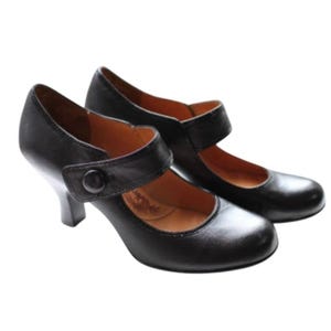 May include: A pair of black leather Mary Jane shoes featuring a rounded toe, a mid-height block heel, and a single strap across the instep secured with a circular button. The interior lining of the shoes is a contrasting tan brown color.