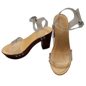 Könnte beinhalten: Ein Paar Plateausandalen mit transparenten Vinylriemen, einem beigen Fußbett und einem braunen Blockabsatz. Das Design umfasst einen transparenten Knöchelriemen mit Metallschnalle und silberfarbene Nieten an der Seite der Sohle. Der Markenname ist im Fersenbereich des Fußbetts aufgedruckt.